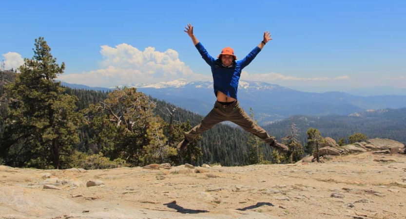 A person stands on an overlook and leaps into the air, raising their arms. Behind them are vast mountains of the high sierra. 